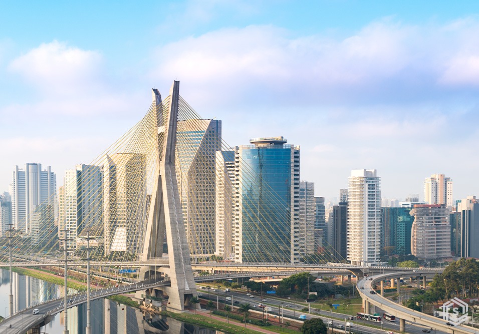 Vista panorâmica de são paulo com destaque para uma ponte estaiada e arranha-céus de diferentes formatos e tamanhos ao fundo, sob céu azul com nuvens.