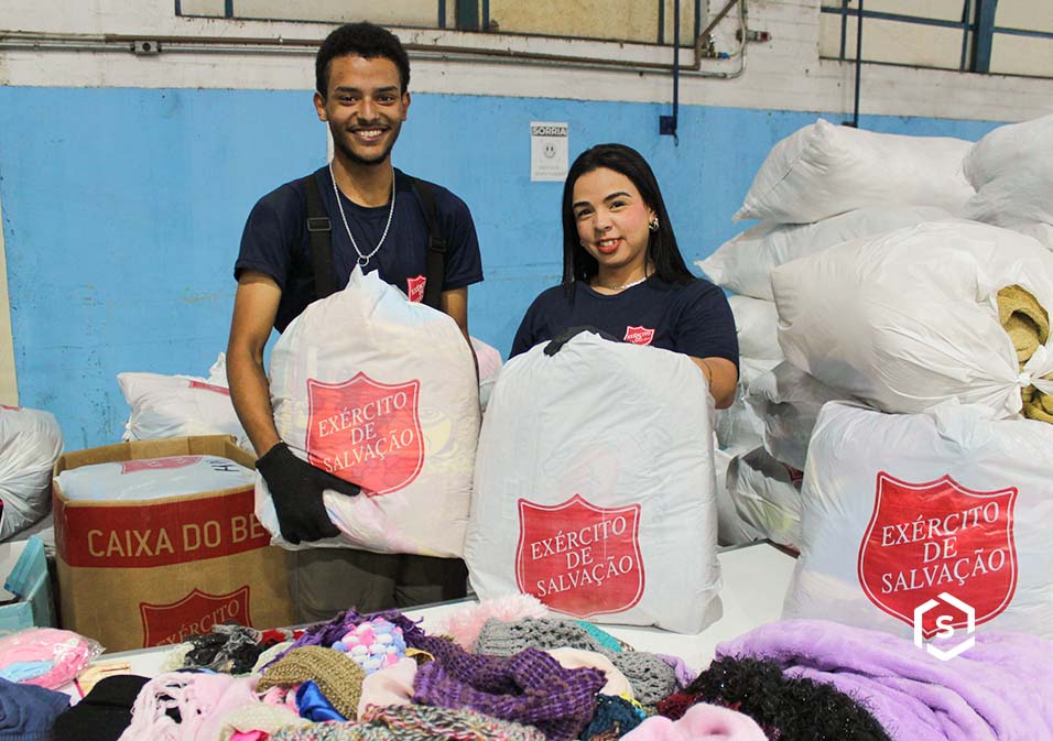 Voluntários do Exército de Salvação do Brasil doando roupas e artigos de higiene em uma campanha de ajuda social, com sorrisos e ambiente interno.