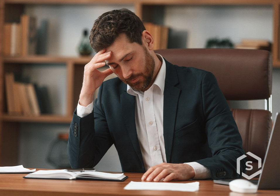 Homem de terno e camisa branca com expressão de preocupação sentado em uma mesa de escritório com livros e computador ao fundo. Homem de terno e camisa branca com expressão de preocupação sentado em uma mesa de escritório com livros e computador ao fundo.