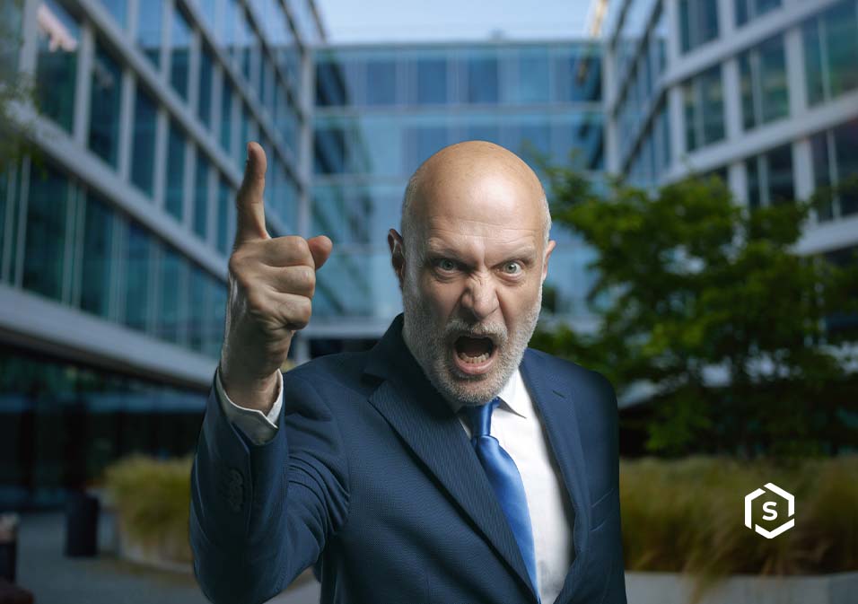 Homem de aparência séria e irritada, vestindo terno azul, gesticulando com dedo levantado na frente de prédio de escritórios modernos Homem de aparência séria e irritada, vestindo terno azul, gesticulando com dedo levantado na frente de prédio de escritórios modernos