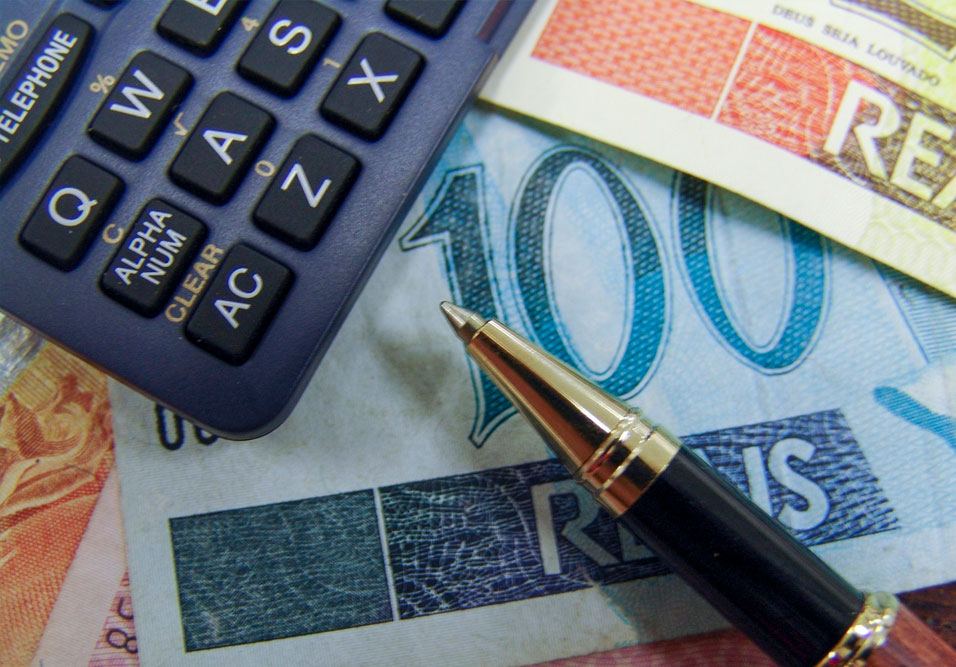 Imagem de uma calculadora, caneta, notas de dinheiro de diferentes denominações e cores, e uma folha de papel em plano de fundo, simbolizando finanças