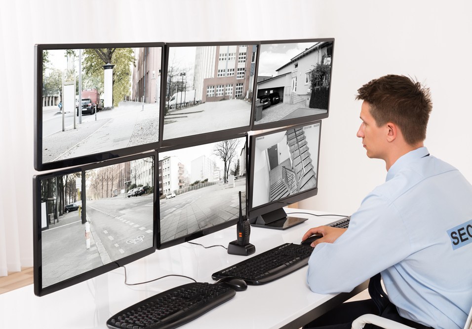Profissional de segurança monitorando câmeras de vigilância em sistema de CFTV com seis telas em guarita de condomínio