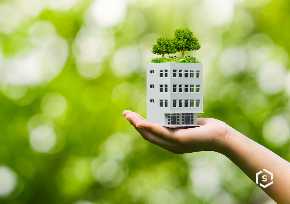 Imagem de uma miniatura de prédio com árvores no topo, simbolizando sustentabilidade e urbanismo verde, sustentada por uma mão, com fundo de natureza.