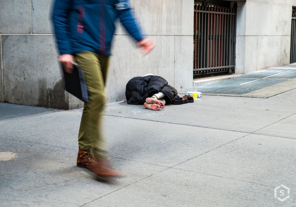 Pessoa morando na rua ao lado de uma parede de prédio, com roupas e pertences ao redor, em uma área urbana e sem abrigo.