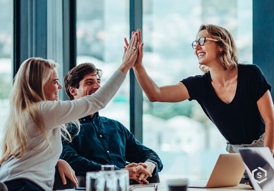 Mulher sorridente dando um high five em um ambiente corporativo com duas pessoas ao seu lado, todos felizes e colaborando em um escritório moderno.