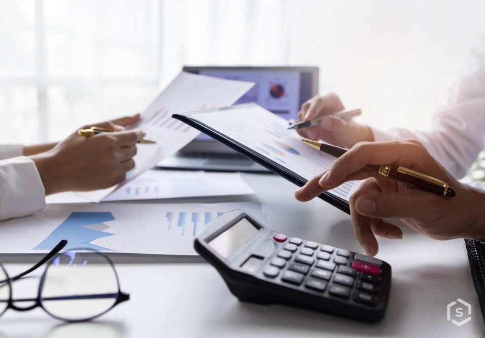 Reunião de pessoas analisando relatórios financeiros com planilhas, tablet, calculadora e laptops em ambiente de escritório bem iluminado.