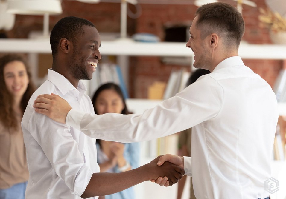 Homem negro e homem branco sorridentes e se cumprimentando em um ambiente de escritório, com colegas ao fundo, celebrando parceria ou sucesso.