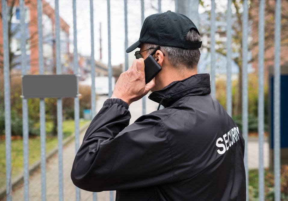 Segurança conversando ao telefone na entrada de uma área com grades, para proteção do condomínio