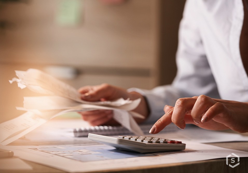Pessoa usando calculadora e papéis em ambiente de escritório para controle financeiro ou cálculo de orçamento.