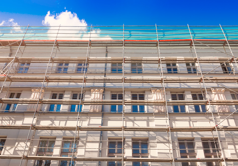 Edifício em construção com escadas de andaime em frente, sob céu azul com nuvens brancas, destacando a fase de reformas na cidade.