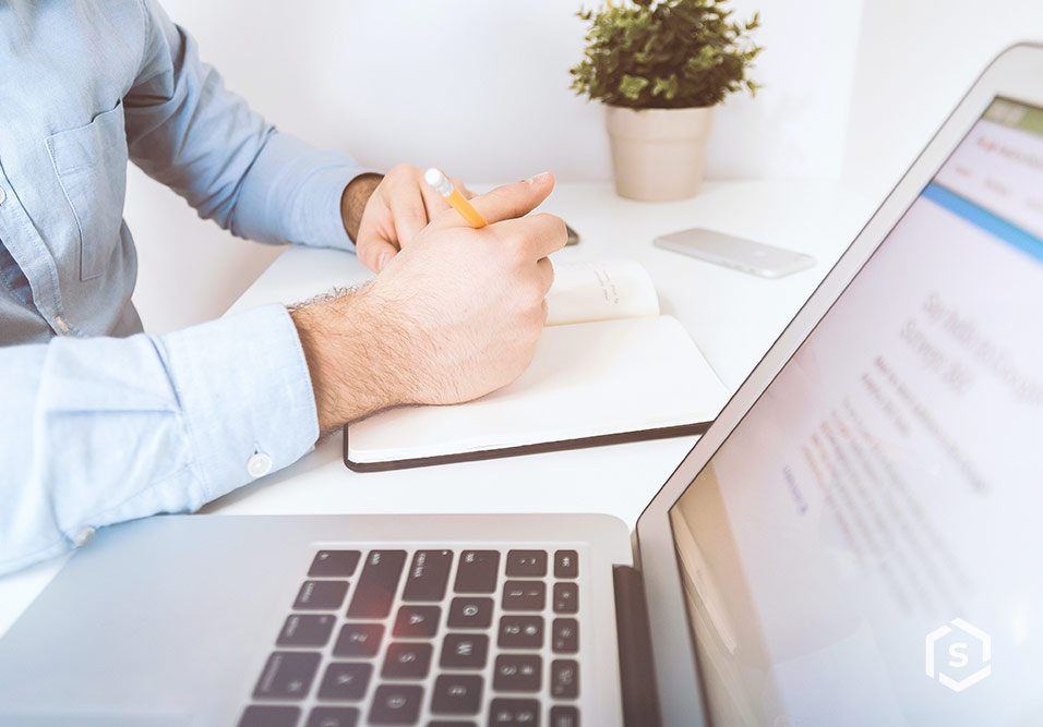 Pessoa anotando informações em um caderno ao lado de um laptop, com planta decorativa ao fundo, ambiente de escritório organizado e luminoso.