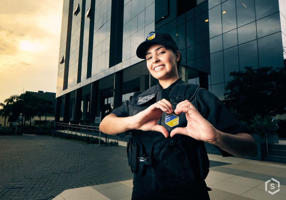 Segurança privada feminina sorridente fazendo coração com as mãos na frente de edifício moderno ao entardecer, promovendo segurança e proteção.