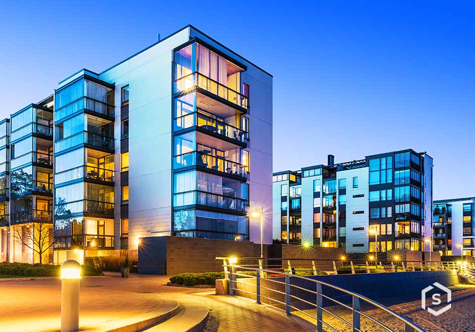 Imagem de um condomínio contemporâneo com múltiplos edifícios de apartamentos residenciais, iluminados à noite, com janelas de vidro e áreas externas 