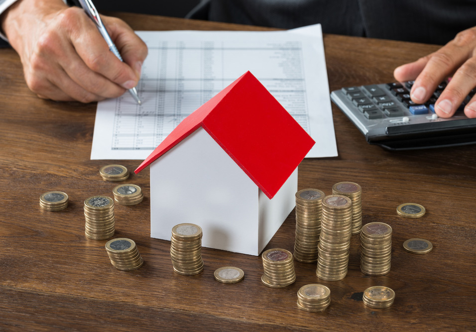 Pessoa assinando documentos enquanto há pilhas de moedas sobre a mesa, com uma maquete de casa com telhado vermelho indicando investimento imobiliário