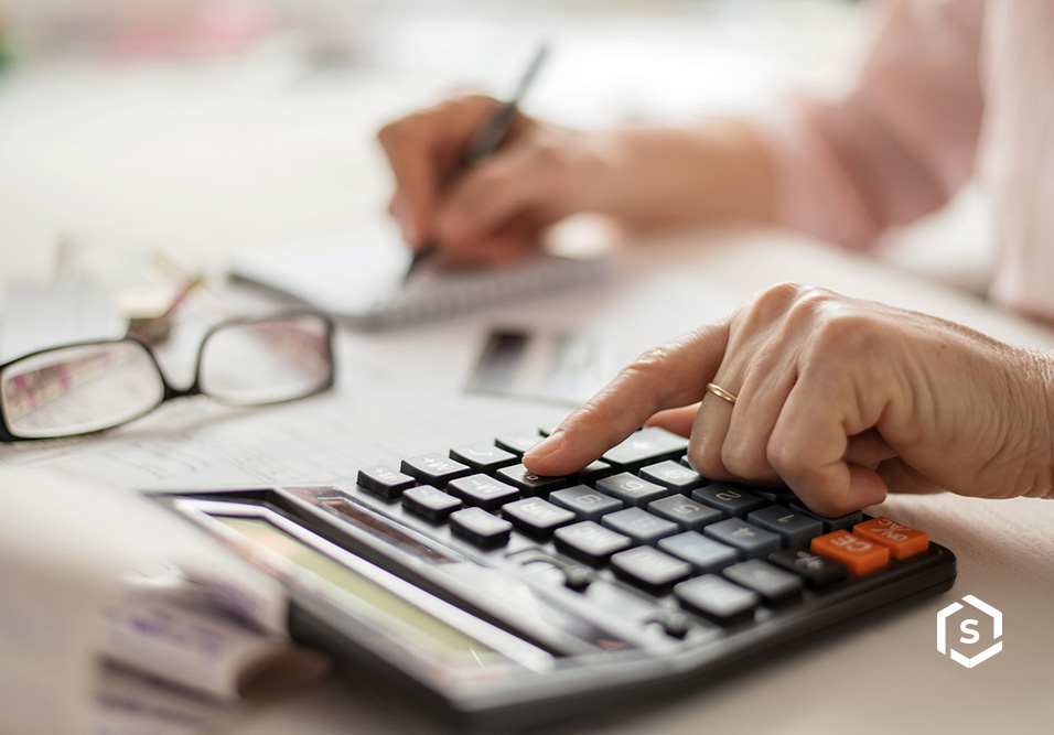 Pessoa utilizando um teclado de computador em uma mesa de escritório, digitando com o dedo e com calculadora, óculos e documentos ao fundo
