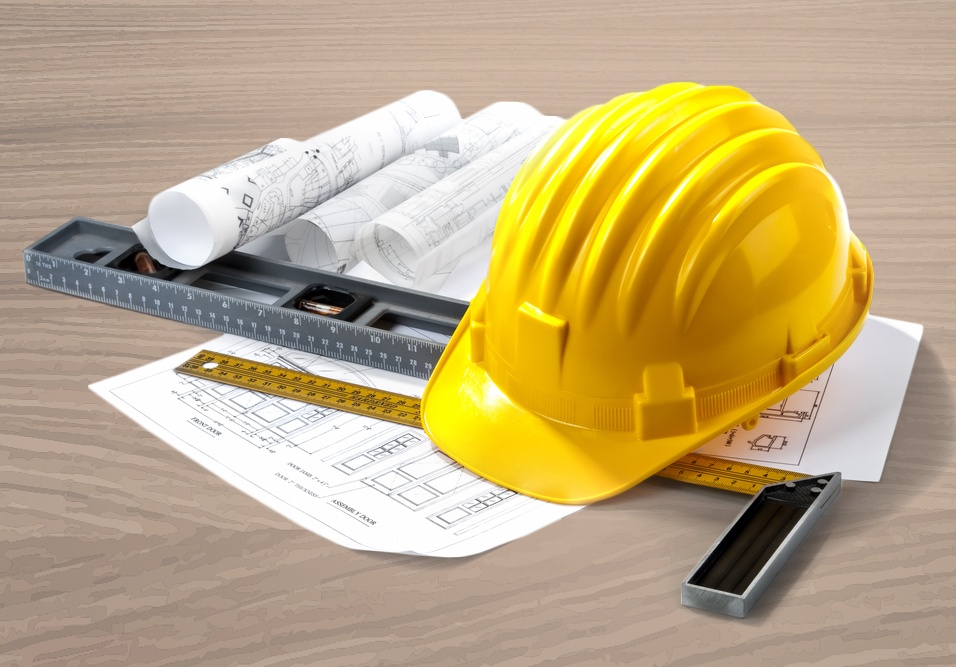 Capacete de segurança amarelo sobre plantas e desenhos técnicos arquitetônicos, ao lado de régua e esquadros de medição em uma mesa de trabalho