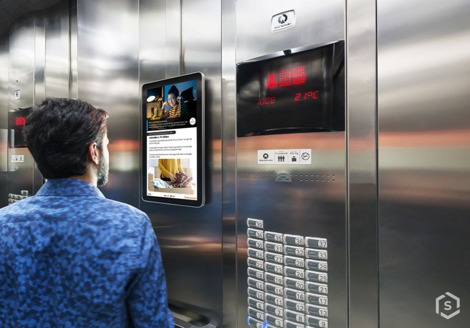 Homem de costas observa uma tela digital em um elevador moderno com telas de informações, botões iluminados e mostrador de andar, em ambiente urbano