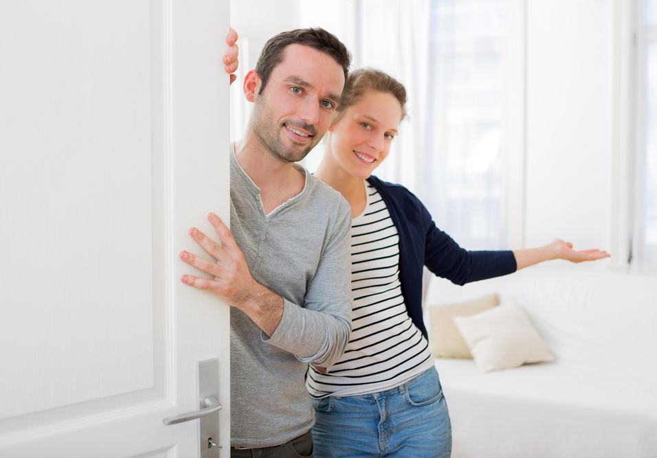 Casal jovem em casa ao lado de uma porta branca, sorrindo e fazendo “toque de parede” com as mãos, em clima de carinho e aconchego no ambiente interno