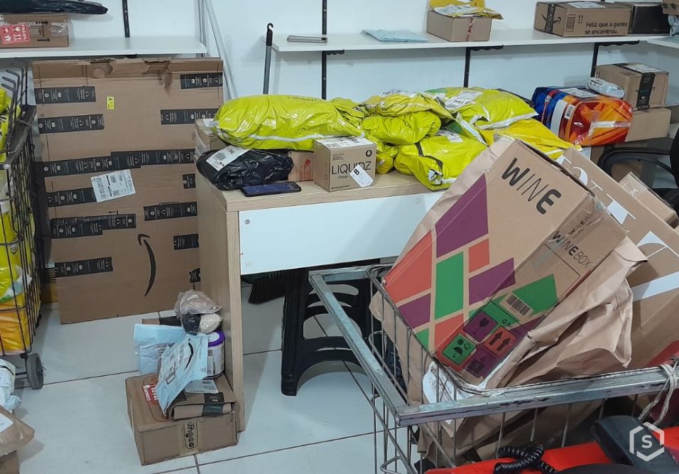Sala com caixas e materiais embalados em prateleiras e carrinho metálico, incluindo caixas de papelão, sacos plásticos e itens diversos