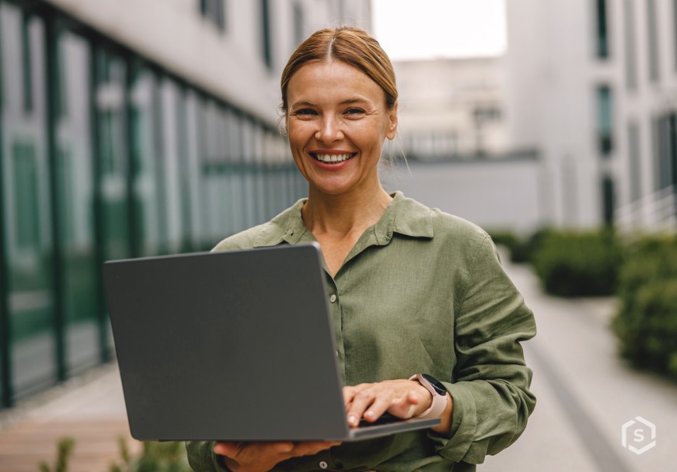 Mulher sorridente em ambiente corporativo externo segura um laptop fechado, usando camisa verde e relógio, em corredor com prédios ao fundo.