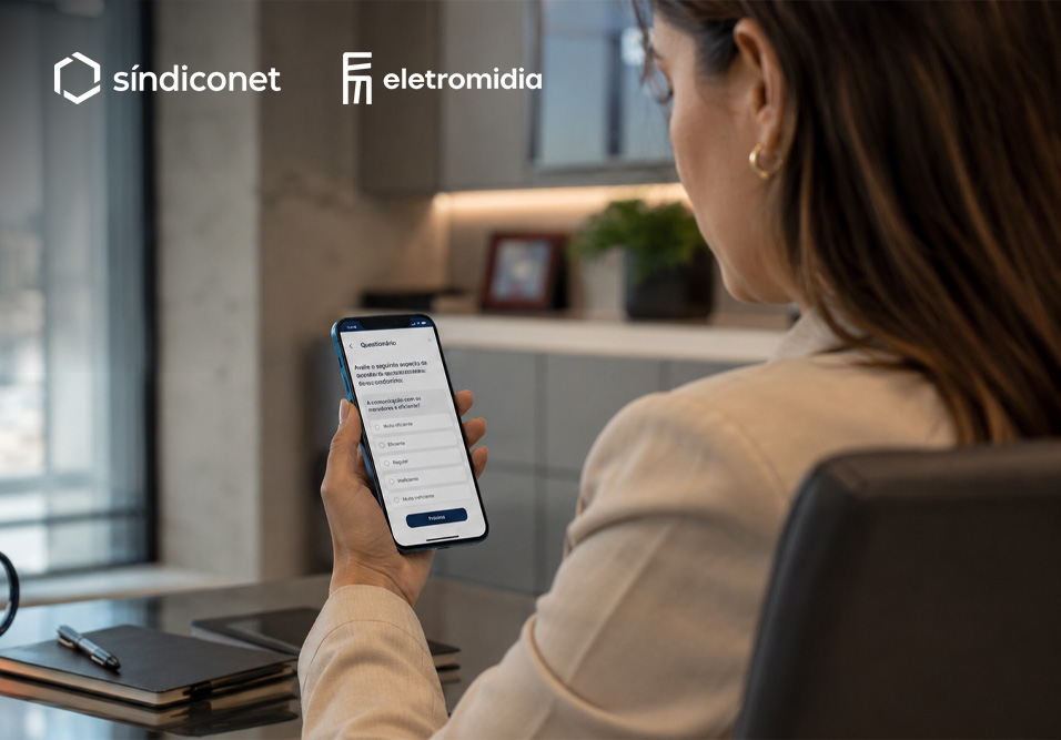 Pessoa segurando um celular com aplicativo de questionário aberto, em ambiente de escritório moderno. Marcas Sindiconet e Eletromidia aparecem no topo