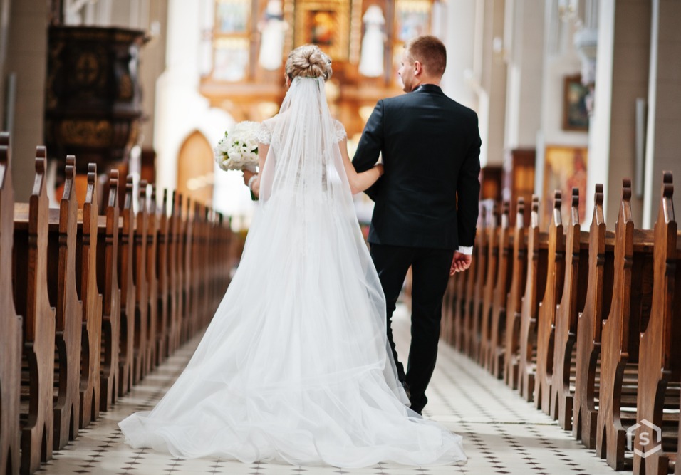 No corredor, uma noiva de vestido branco longo com véu e buquê caminha de mãos dadas com o noivo de terno, durante a cerimônia.