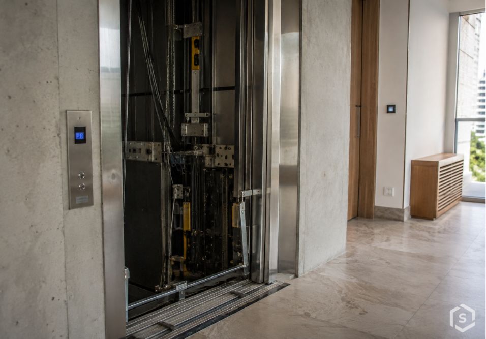 Corredor interno de prédio com elevador ao fundo, portas de vidro e estrutura metálica do elevador, piso de mármore e paredes claras