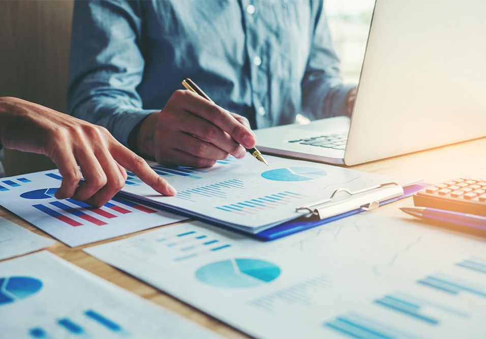 Síndico fazendo anotações e analisando gráficos e relatórios financeiros, com notebook ao fundo e documentos de dados sobre a mesa.