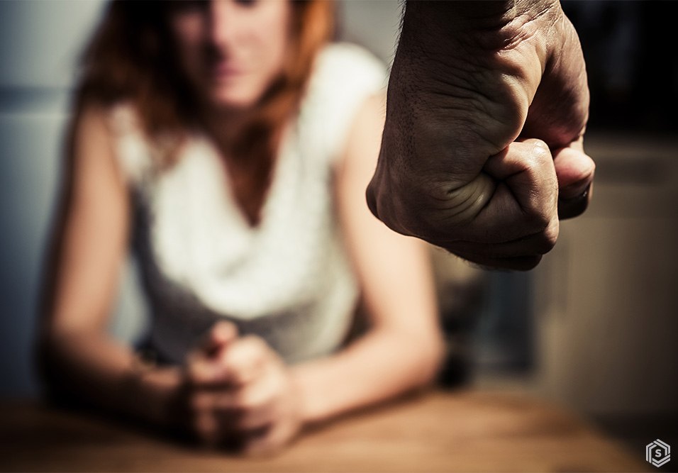 Imagem em close de uma mão masculina cerrada em gesto de ameaça, enquanto uma mulher ao fundo aparece sentada com expressão de preocupação