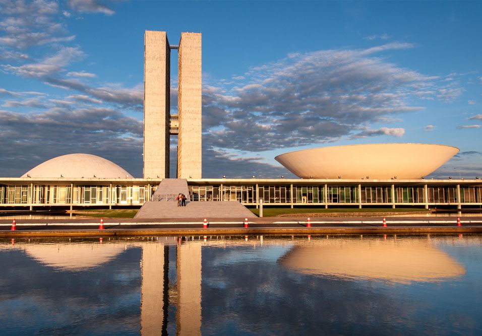 Vista externa do Congresso Nacional com espelhos d’água refletindo o prédio e o céu azul ao fundo, em uma cena ao ar livre