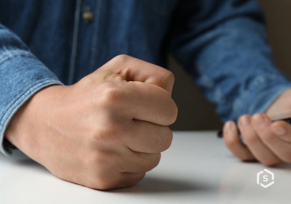 Pessoa com uma mão serrada e a outra usando smartphone, digitando, roupa casual de jeans, foco na mão e no aparelho, cenário neutro.